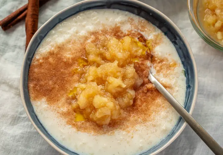 Cremiger Milchreis mit Apfelmus und Zimt, der weltbester Milchreis-Geschmack verspricht. Daneben liegen Zimtstangen.