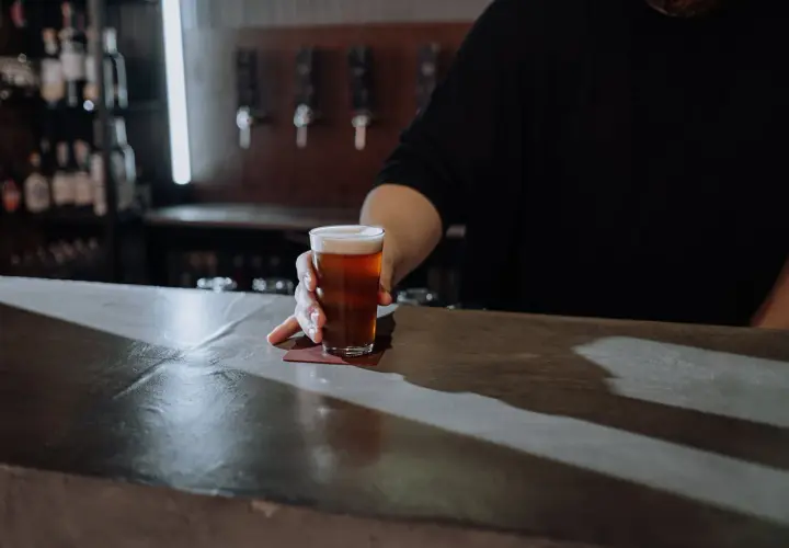 Ein Glas Bier wird über den Tresen geschoben. Der Unterschied zwischen Bier und Pils ist hier nicht ersichtlich, aber die Farbe des Bieres ist bernsteinfarben.