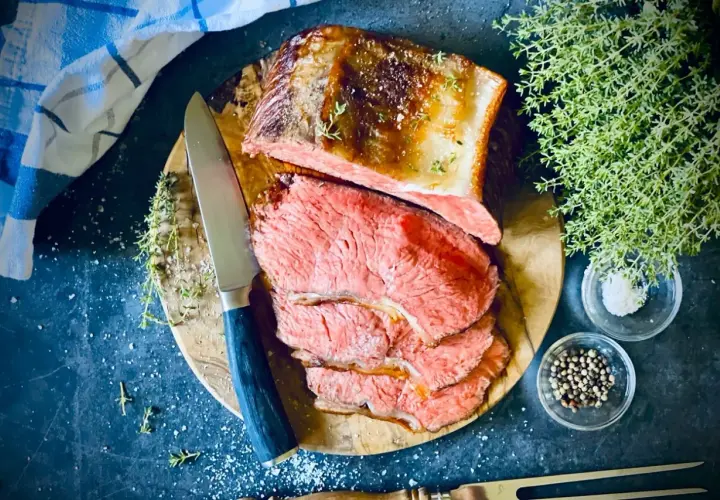 Saftiger Rostbraten, perfekt im Ofen zubereitet, liegt auf einem Holzbrett. Daneben liegen Kräuter, Salz und Pfefferkörner.