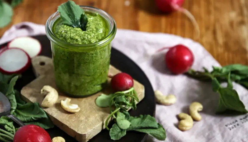 Frisches Radieschen Pesto in einem Glas, umgeben von Radieschen, Cashewkernen und grünen Blättern auf einem Holztisch.