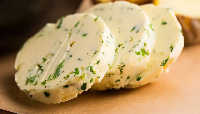 Drei Scheiben Kapuzinerkressebutter liegen auf einem braunen Untergrund. Die Butter ist mit grünen Kräutern durchsetzt.