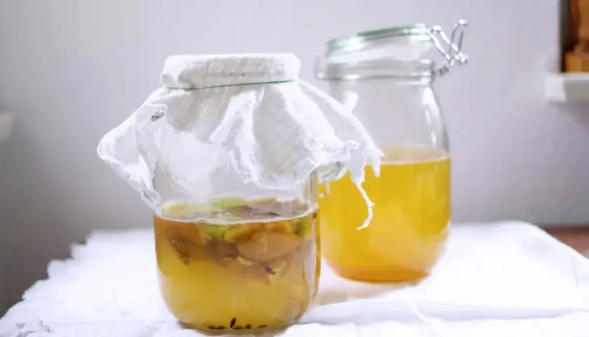 Zwei Gläser mit gelblicher Flüssigkeit. Im vorderen Glas schwimmen Apfelstücke unter einem weißen Tuch. Hier wird Apfelessig selber gemacht.