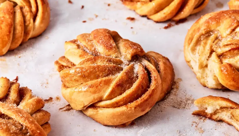 Goldbraune Kardamomschnecken, bestreut mit Zucker und Zimt, liegen auf Backpapier.