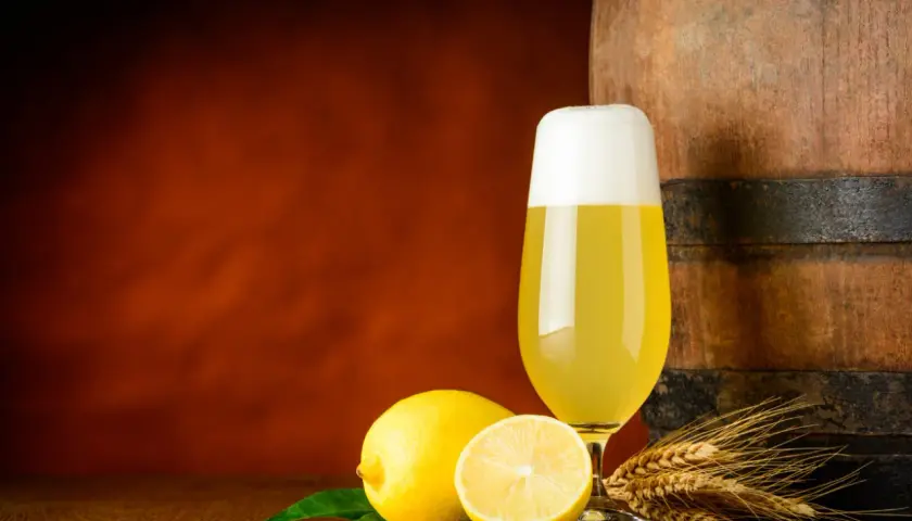 Ein Glas Radler steht neben Zitronen und Gerstenähren vor einem Holzfass.