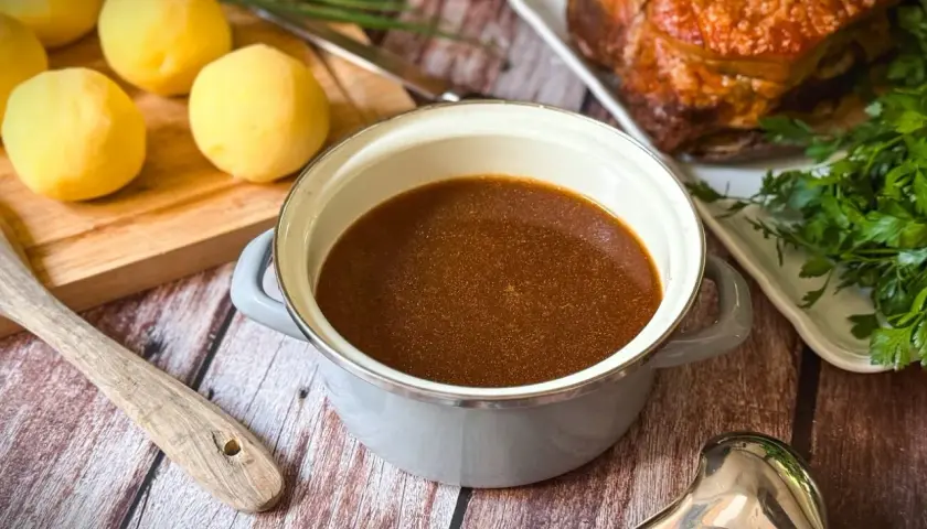 Ein Topf mit köstlichem Bratenfond, bereit, um das Gericht zu verfeinern. Daneben liegen gekochte Kartoffeln und ein Stück Braten.
