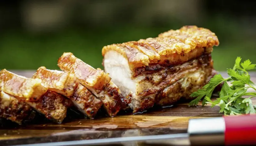 Ein perfekt gebratener, knuspriger Schweinebauch mit goldbrauner Kruste, auf einem Holzbrett mit Petersilie und einem Messer.
