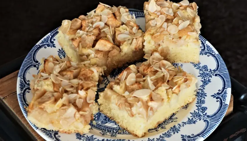 Vier Stücke saftiger Apfelkuchen vom Blech mit Mandeln, auf einem blauen Teller angerichtet.