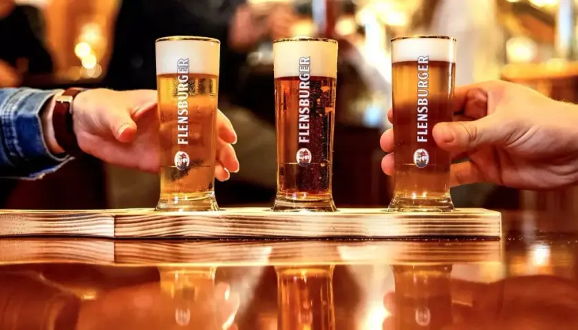 Drei Gläser Flensburger Bier mit cremigem **Bier schaum** stehen auf einem Holztablett. Hände greifen nach den Gläsern.