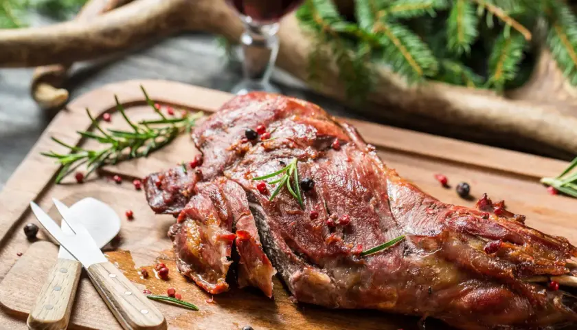 Ein saftiger Rehschlegel, perfekt gegrillt, liegt auf einem Holzbrett. Gewürze und Rosmarin zieren das Fleisch. Ein Glas Rotwein und Hirschgeweih im Hintergrund deuten auf ein festliches rehschlegel rezept hin.