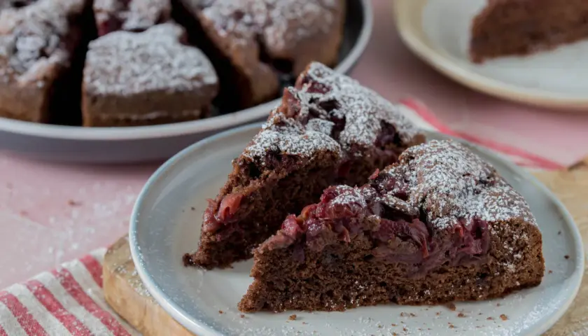 Saftiger Schoko-Kirsch-Kuchen aus der Springform, mit Puderzucker bestäubt. Zwei Stücke auf einem Teller, der ganze Kuchen im Hintergrund.