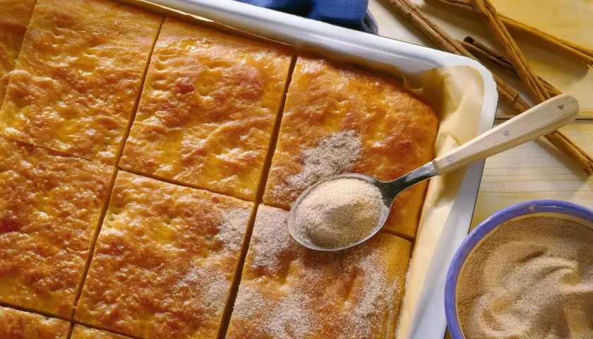 Goldbrauner Kartoffelkuchen vom Blech, frisch aus dem Ofen, wird mit Zimt-Zucker bestreut. Daneben liegen Zimtstangen und eine Schüssel mit der süßen Mischung.
