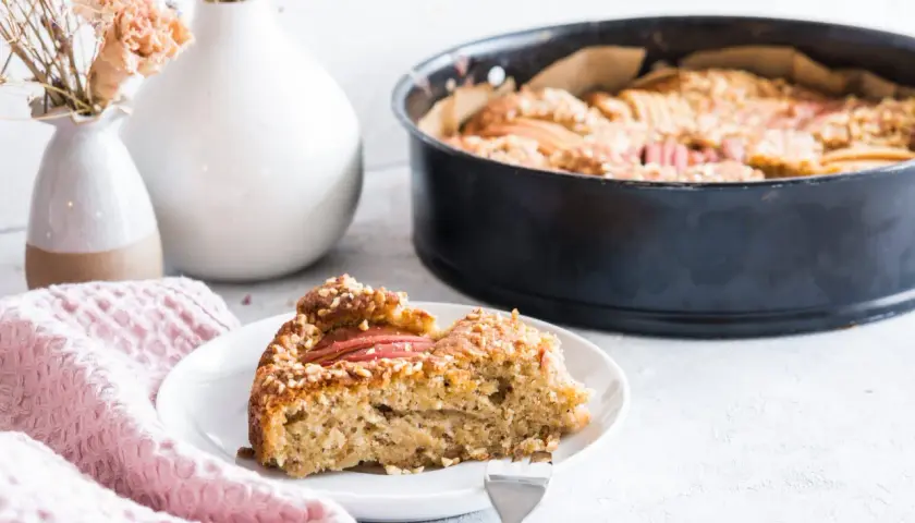 Ein Stück Apfelkuchen mit Haselnuss-Streuseln auf einem Teller, daneben eine Backform mit dem restlichen Kuchen.