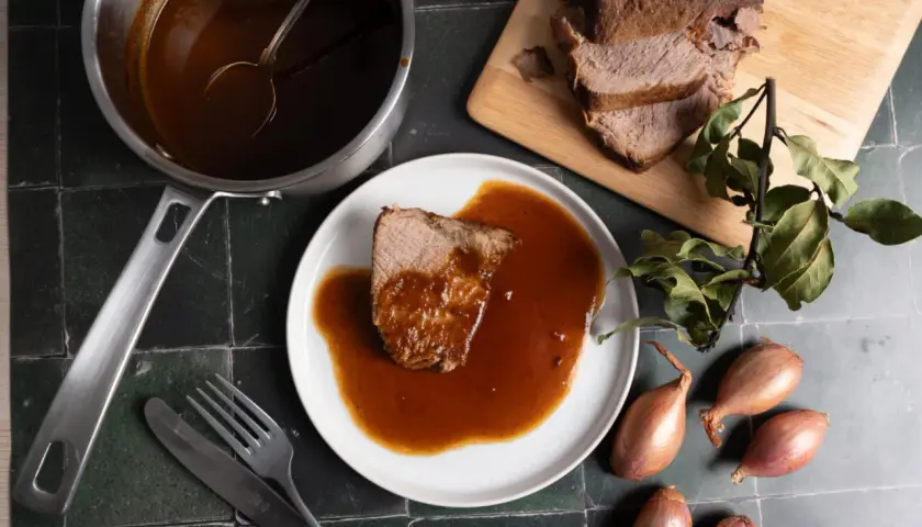 Ein Stück Rinderbraten mit aromatischer Rinderbraten Sauce auf einem Teller, daneben ein Topf mit Sauce, ein Messer und eine Gabel.