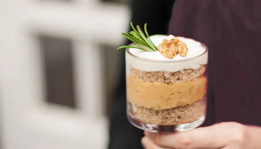 Ein Glas mit Schichten von Kuchen und cremiger Karamell-Mousse, garniert mit Rosmarin und einer Walnusshälfte.