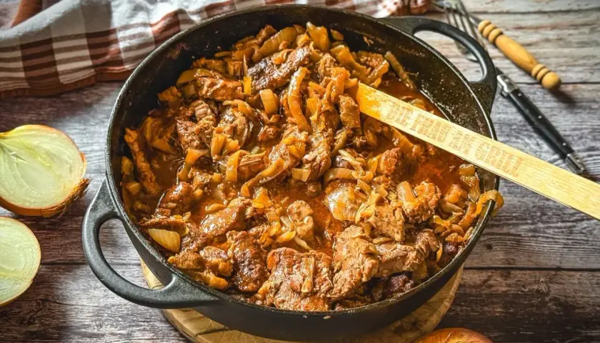 Saftiger Zwiebelbraten in einer Gusseisenpfanne mit Zwiebeln und Fleisch, daneben halbierte Zwiebeln.