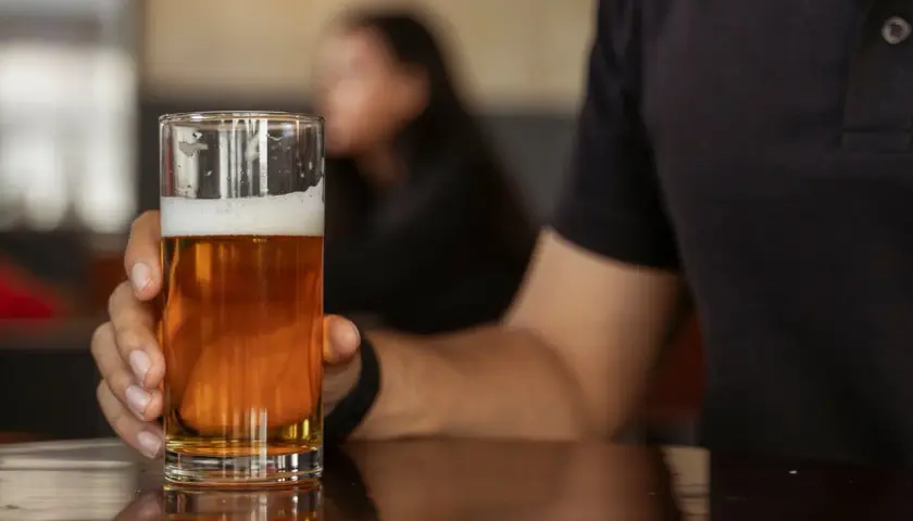 Mann hält ein Glas mit hellem englischem Bier. Der Schaum krönt das goldene Getränk, während im Hintergrund eine Person verschwommen zu sehen ist.