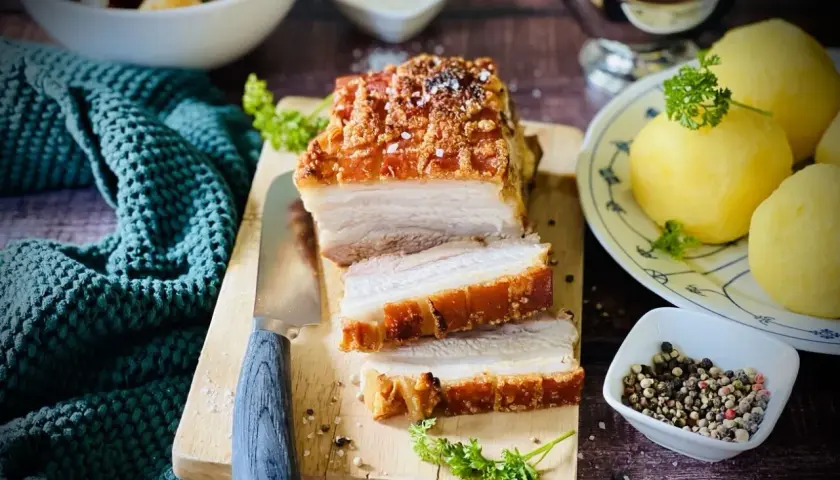 Ein saftiger Schweinebraten mit knuspriger Kruste, auf einem Holzbrett angeschnitten, daneben Klöße und Soße.