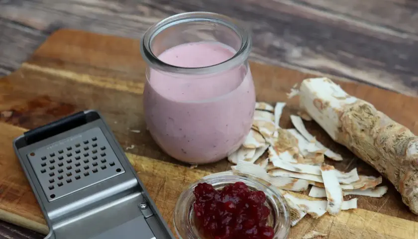 Meerrettichschmand in einem Glas, daneben frisch geriebener Meerrettich, eine Reibe und Preiselbeeren.