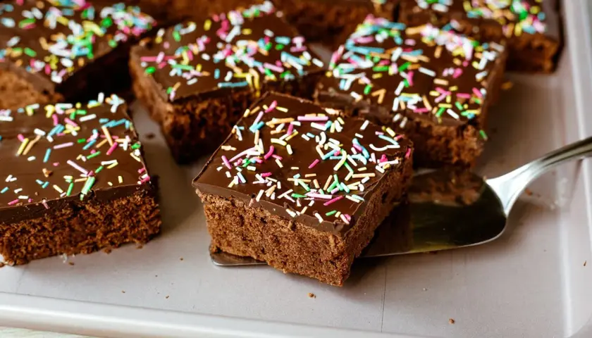 Saftiger schokokuchen blech mit bunten Streuseln, bereit zum Servieren. Ein Stück wird gerade mit einem Tortenheber herausgenommen.