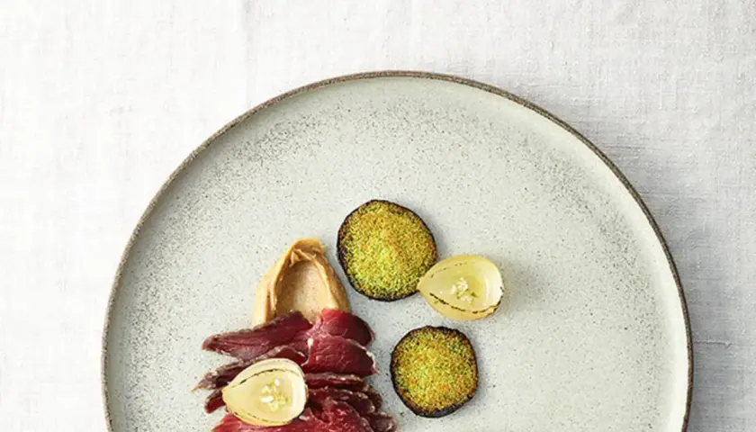 Gebeiztes Rinderfilet mit cremiger Beilage, karamellisierten Zwiebeln und grünen, knusprigen Scheiben auf einem Teller.