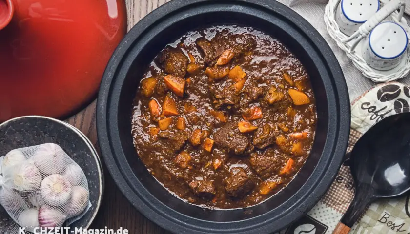 Herzhaftes Rehhals Rezept in einem Schmortopf, mit Karottenstücken und Knoblauch daneben.