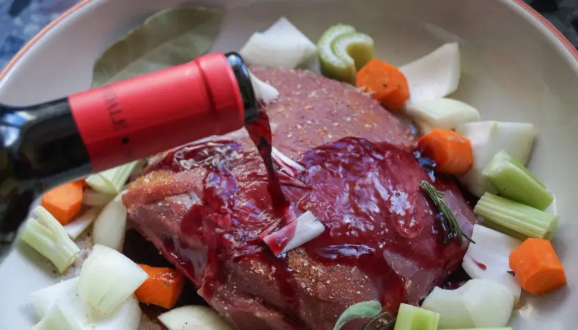 Rinderbraten in Rotwein einlegen. Ein roter Weinstrahl ergießt sich über das Fleisch, umgeben von Karotten, Sellerie und Zwiebeln.