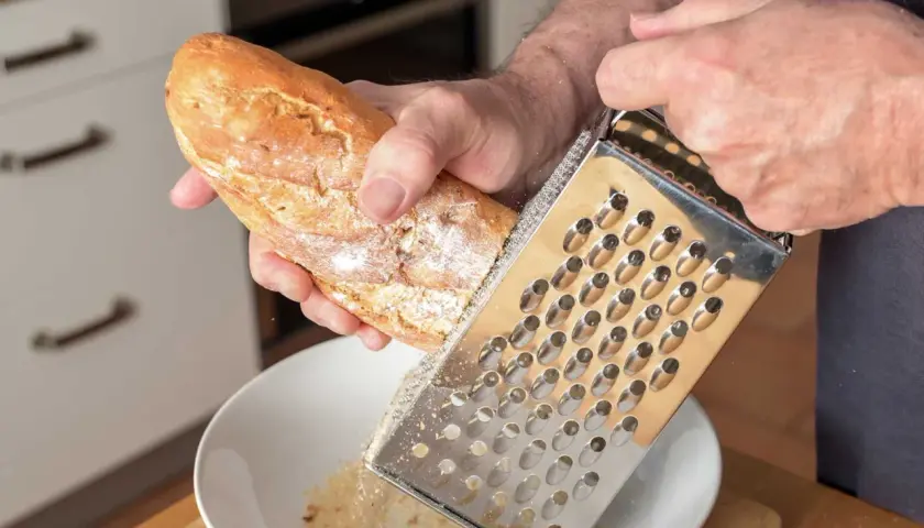 Mann reibt ein Baguette über einer Schüssel, um Paniermehl selber zu machen. Frische Brösel für die Küche.