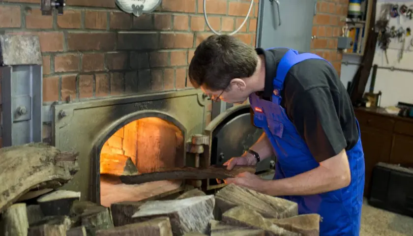 Mann legt Holz in den Ofen für die Rauchbier Herstellung. Das Feuer glüht hell.