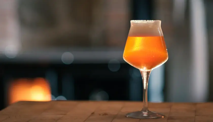 Ein Glas mit bernsteinfarbenem Bier, das nach pale ale malz duftet, steht auf einem Holztisch. Im Hintergrund glüht ein Kaminfeuer.