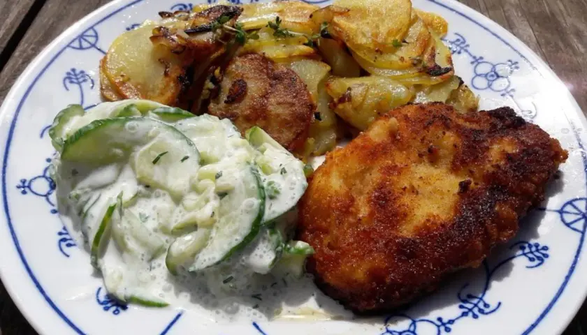 Ein knuspriges Schnitzel mit goldbraunen Ofenkartoffeln und einem cremigen Gurkensalat auf einem blauen Zwiebelmuster-Teller.