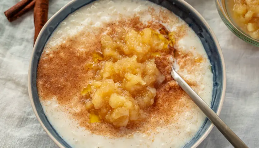 Cremiger Milchreis mit Apfelmus und Zimt, der weltbester Milchreis-Geschmack verspricht. Daneben liegen Zimtstangen.