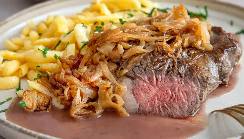 Zwiebelrostbraten im Backofen, saftig rosa gebraten, mit goldbraunen Röstzwiebeln und Spätzle auf einem Teller.