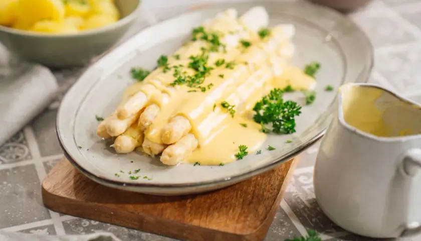 Spargel mit cremiger Sauce Hollandaise, ein Klassiker, der mit diesem Rezept gelingt. Beilagen wie Kartoffeln sind im Hintergrund zu sehen.