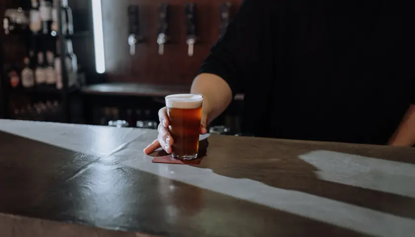 Ein Glas Bier wird über den Tresen geschoben. Der Unterschied zwischen Bier und Pils ist hier nicht ersichtlich, aber die Farbe des Bieres ist bernsteinfarben.