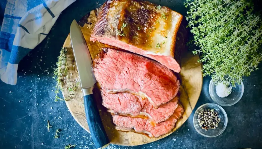 Saftiger Rostbraten, perfekt im Ofen zubereitet, liegt auf einem Holzbrett. Daneben liegen Kräuter, Salz und Pfefferkörner.