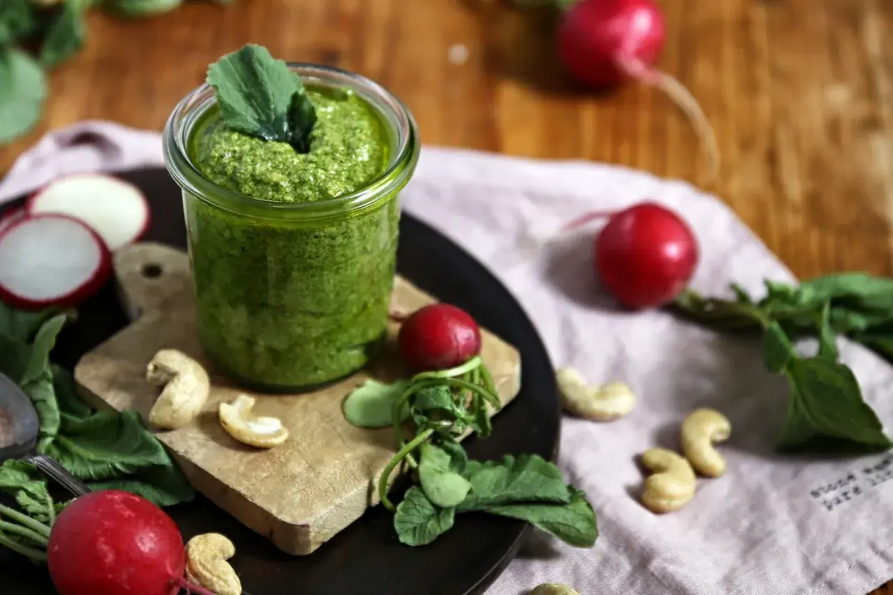 Frisches Radieschen Pesto in einem Glas, umgeben von Radieschen, Cashewkernen und grünen Blättern auf einem Holztisch.