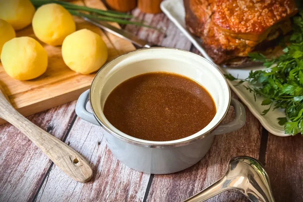 Ein Topf mit köstlichem Bratenfond, bereit, um das Gericht zu verfeinern. Daneben liegen gekochte Kartoffeln und ein Stück Braten.