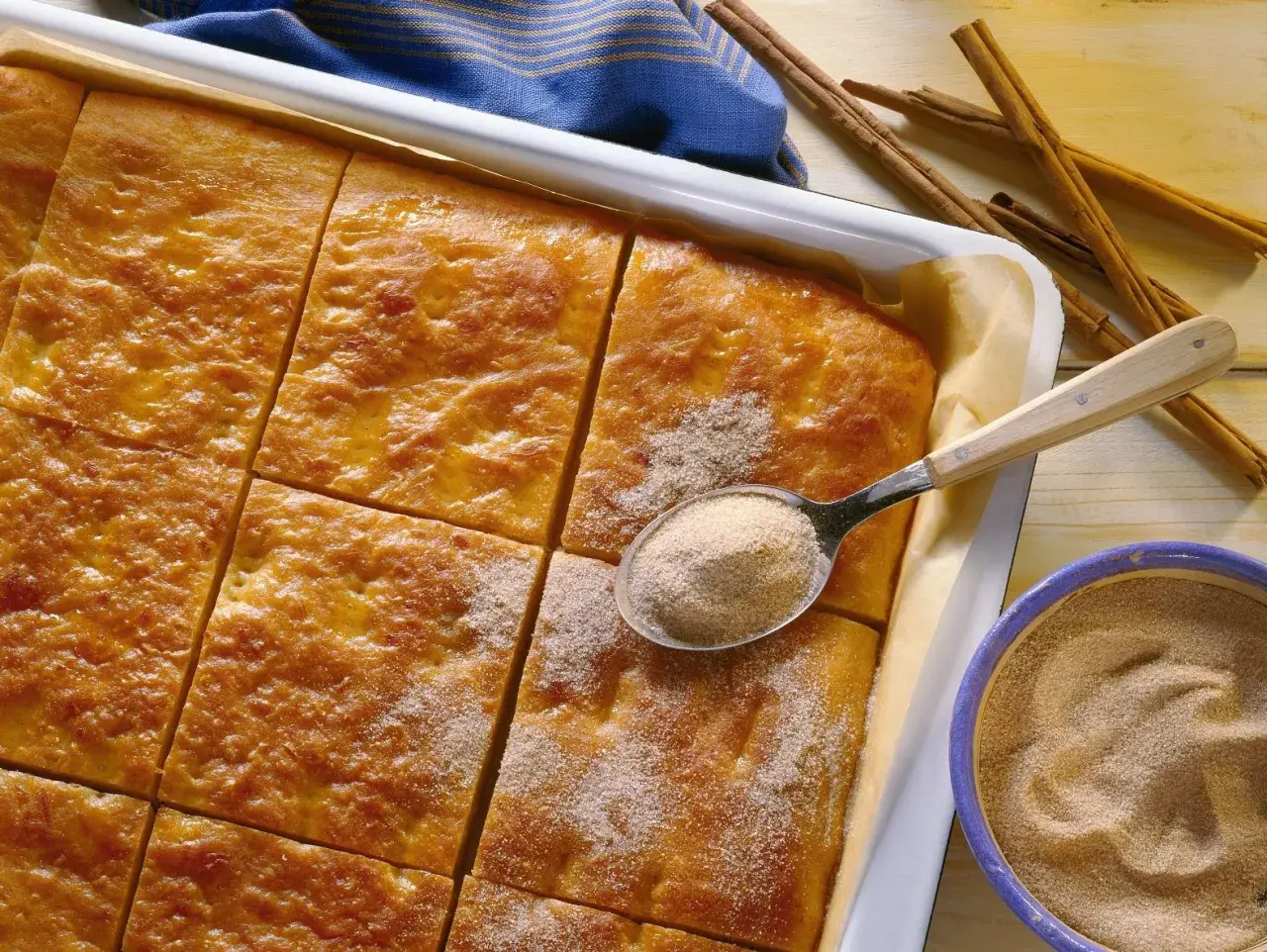 Goldbrauner Kartoffelkuchen vom Blech, frisch aus dem Ofen, wird mit Zimt-Zucker bestreut. Daneben liegen Zimtstangen und eine Schüssel mit der süßen Mischung.