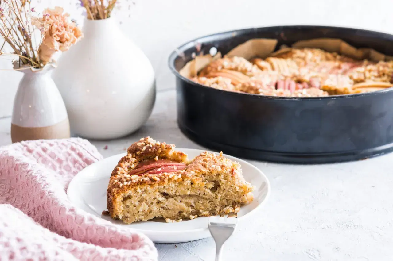 Ein Stück Apfelkuchen mit Haselnuss-Streuseln auf einem Teller, daneben eine Backform mit dem restlichen Kuchen.