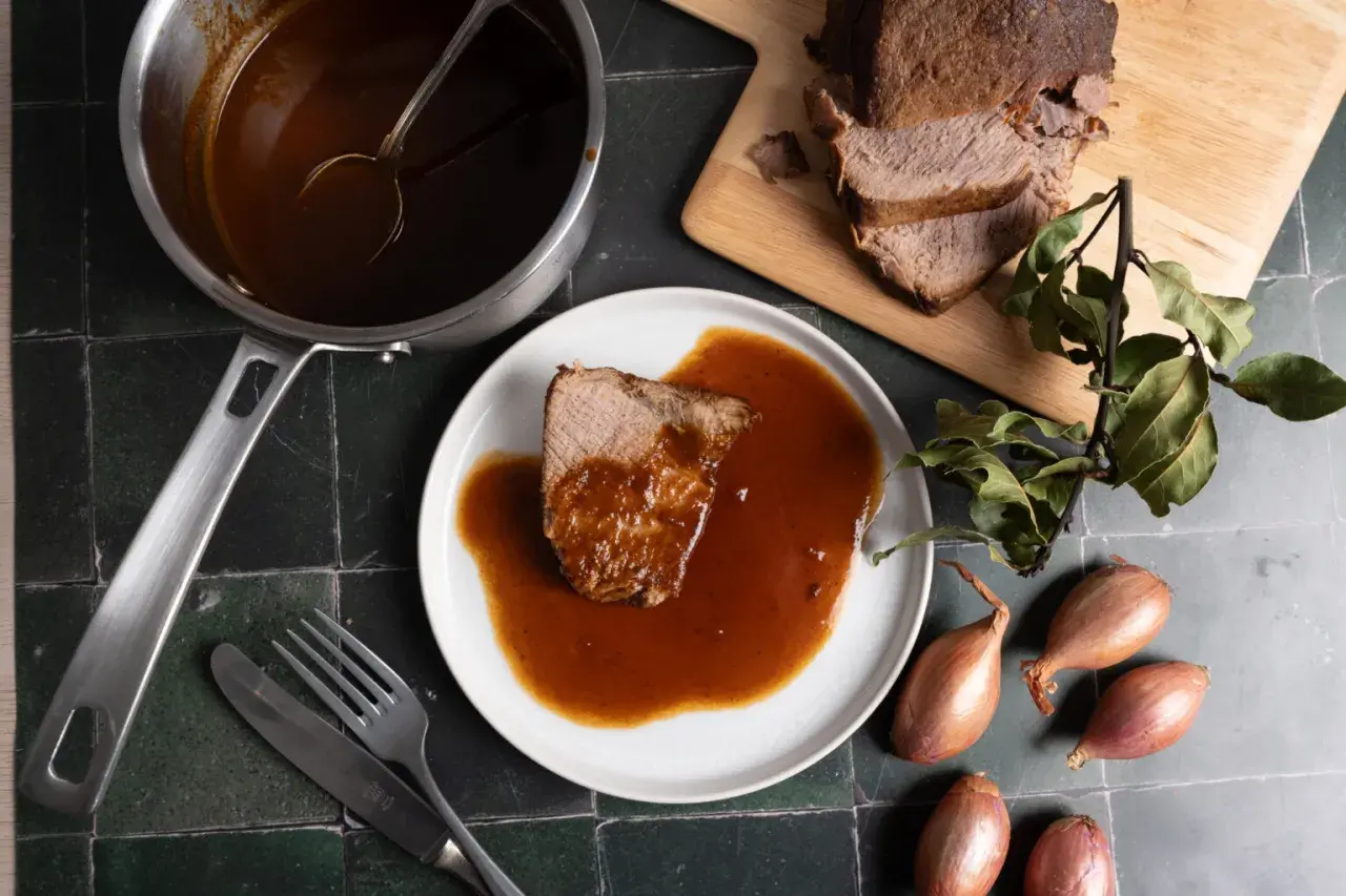 Ein Stück Rinderbraten mit aromatischer Rinderbraten Sauce auf einem Teller, daneben ein Topf mit Sauce, ein Messer und eine Gabel.