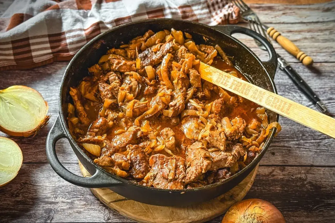 Saftiger Zwiebelbraten in einer Gusseisenpfanne mit Zwiebeln und Fleisch, daneben halbierte Zwiebeln.