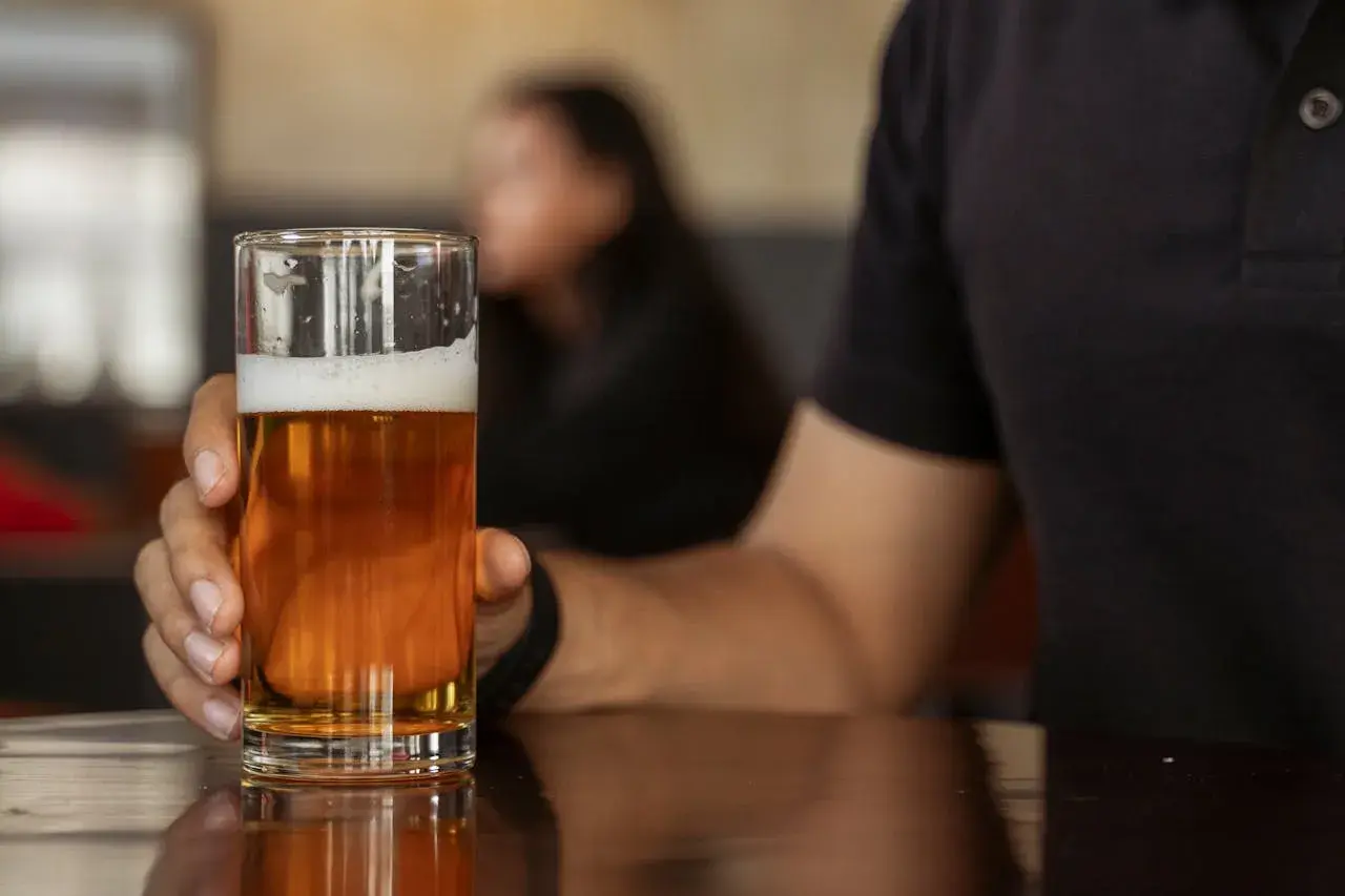 Mann hält ein Glas mit hellem englischem Bier. Der Schaum krönt das goldene Getränk, während im Hintergrund eine Person verschwommen zu sehen ist.