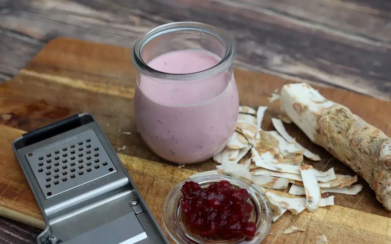 Meerrettichschmand in einem Glas, daneben frisch geriebener Meerrettich, eine Reibe und Preiselbeeren.