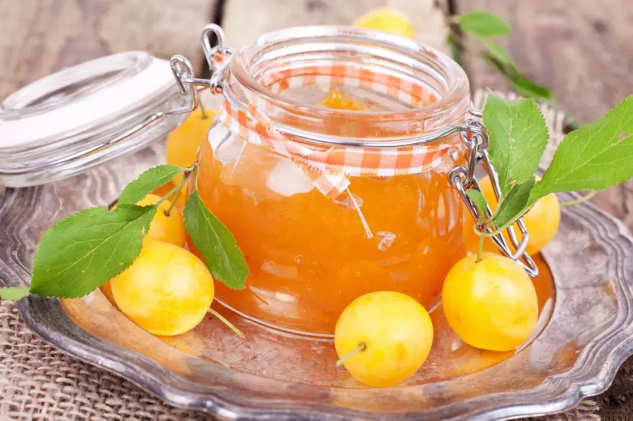 Ein Glas mit leuchtend orangefarbener Mirabellenmarmelade, umgeben von frischen Früchten und Blättern. Ein schönes Bild für ein Mirabellenmarmelade Rezept.