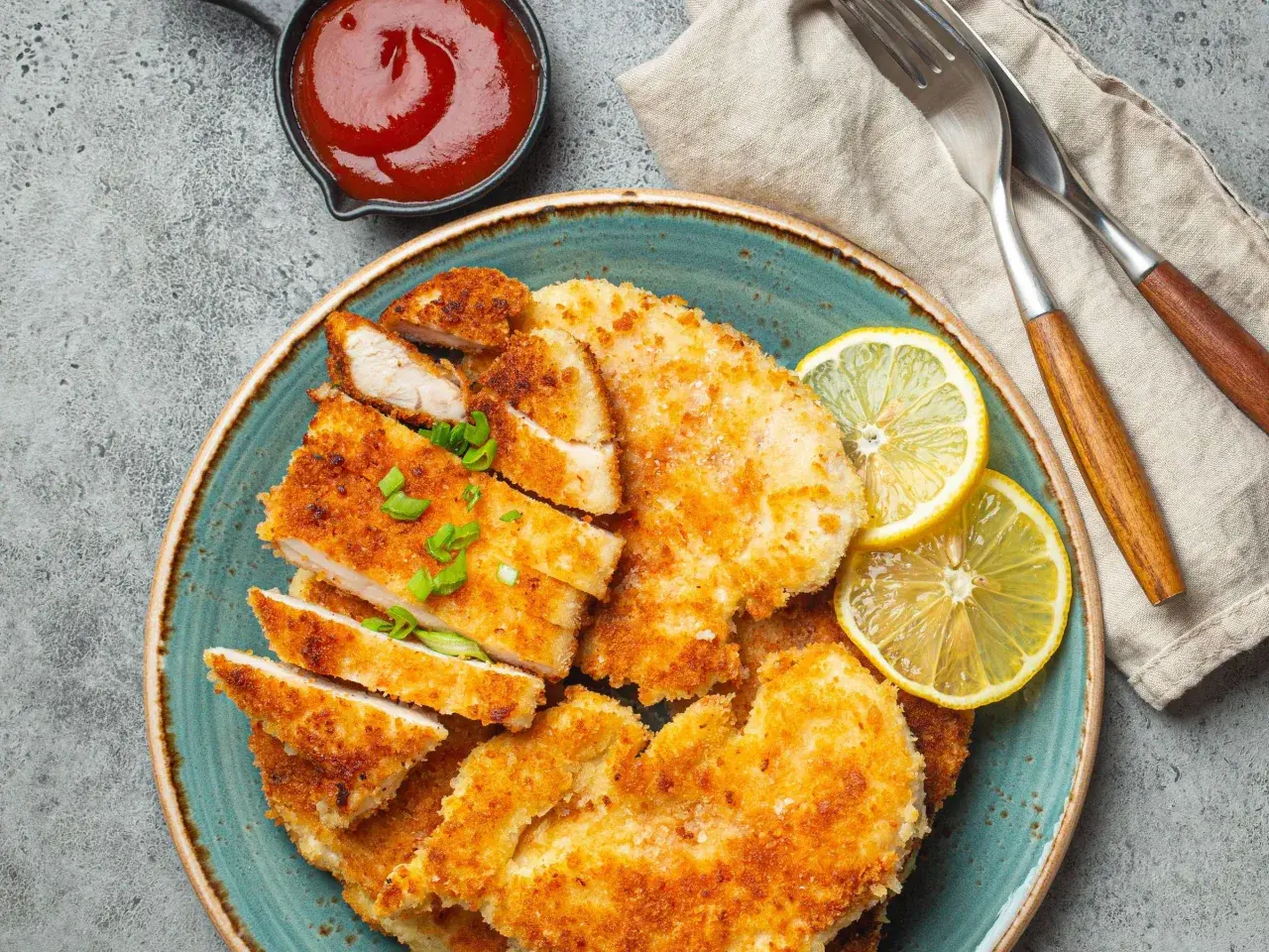 Knusprige Schnitzel mit Treber Panade, garniert mit Zitrone und Schnittlauch, daneben eine Schale Ketchup.