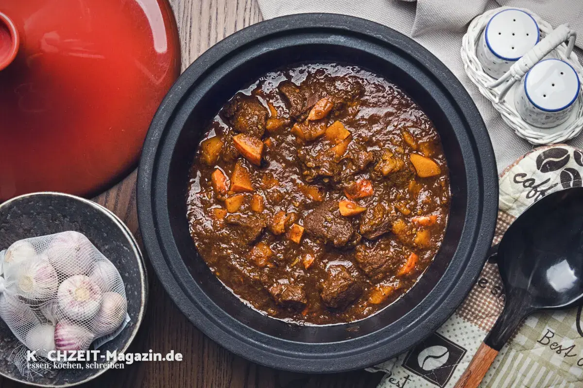 Herzhaftes Rehhals Rezept in einem Schmortopf, mit Karottenstücken und Knoblauch daneben.