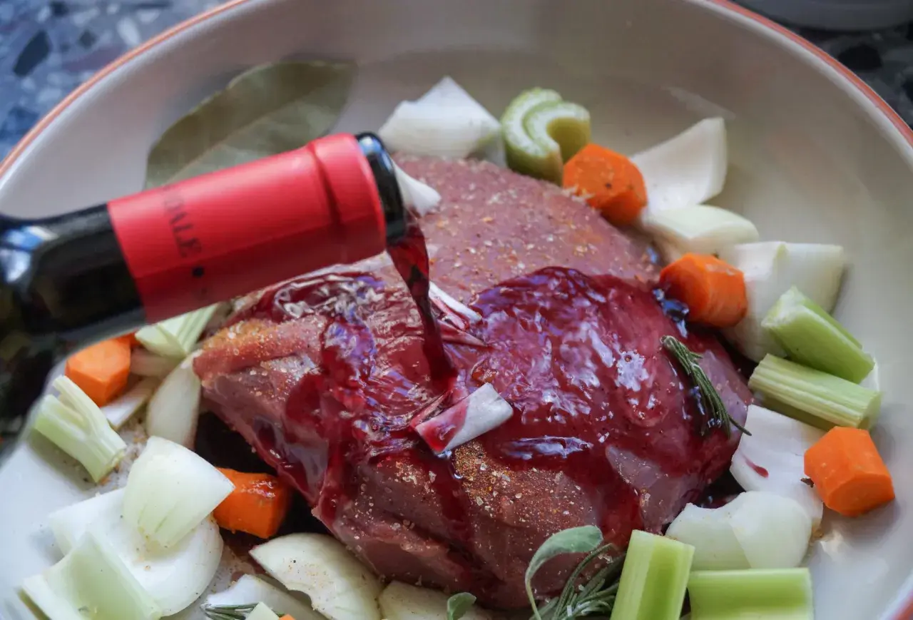 Rinderbraten in Rotwein einlegen. Ein roter Weinstrahl ergießt sich über das Fleisch, umgeben von Karotten, Sellerie und Zwiebeln.
