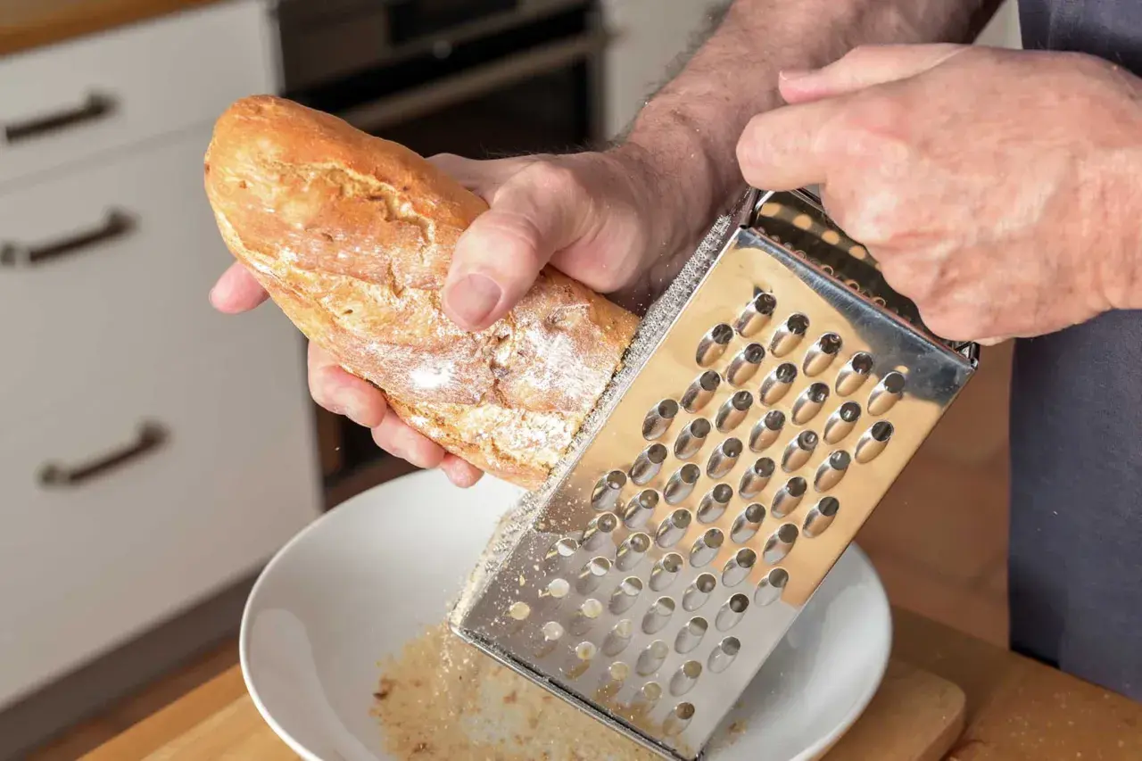 Mann reibt ein Baguette über einer Schüssel, um Paniermehl selber zu machen. Frische Brösel für die Küche.