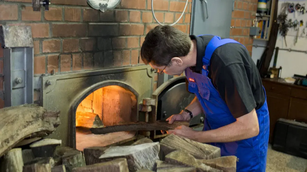 Mann legt Holz in den Ofen für die Rauchbier Herstellung. Das Feuer glüht hell.