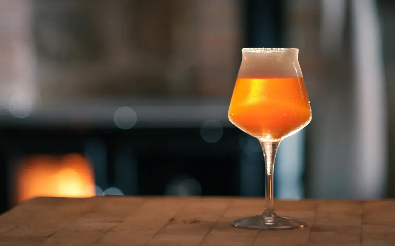 Ein Glas mit bernsteinfarbenem Bier, das nach pale ale malz duftet, steht auf einem Holztisch. Im Hintergrund glüht ein Kaminfeuer.